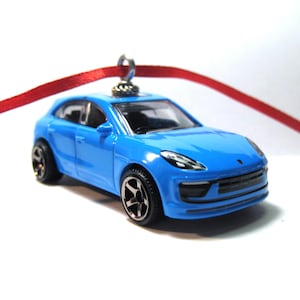 May include: A bright blue toy car with black wheels and a red ribbon for hanging. The miniature vehicle has a detailed design, including headlights and a front grill. A small, silver metal loop is attached to the top of the car.