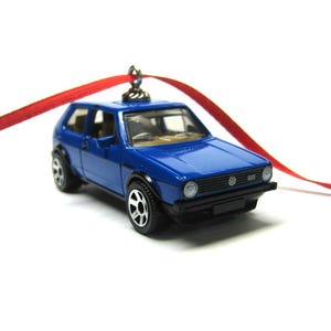 May include: A miniature blue Volkswagen GTI toy car with black wheels and a red ribbon for hanging. The car has a detailed design, including headlights, a front grill, and the GTI logo. The background is white.