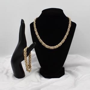 May include: A gold-toned Byzantine chain necklace and a matching Byzantine chain bracelet. The necklace is displayed on a black bust, while the bracelet is displayed on a black hand-shaped stand, both featuring intricate interlocking links.
