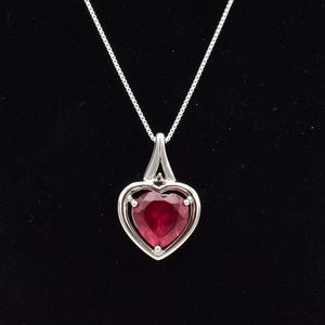 May include: A silver necklace with a heart-shaped pendant. The pendant has a faceted, deep red gemstone set in a silver frame. The chain is a delicate silver box chain, against a black background.
