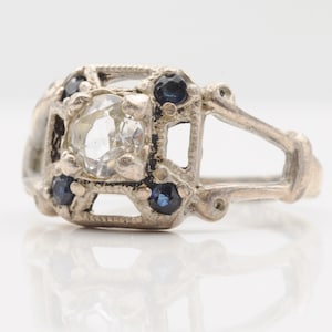 Op de afbeelding: Een zilverkleurige ring met een centrale heldere, gefacetteerde steen omringd door vier kleine, donkerblauwe, rond geslepen stenen. De metalen zetting heeft een open, geometrisch en getextureerd ontwerp met een gespleten ringband aan de achterzijde.