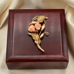 May include: A gold-toned floral brooch with two coral-colored rose blossoms and detailed leaves, set on a dark wooden box. The brooch is a detailed piece of jewelry, likely vintage.