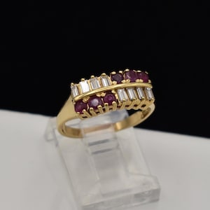 May include: A gold ring featuring two parallel rows of baguette-cut clear stones separated by a central row of round-cut deep red rubies. The band is crafted from polished yellow gold, showcasing a structured design with alternating gemstone settings.