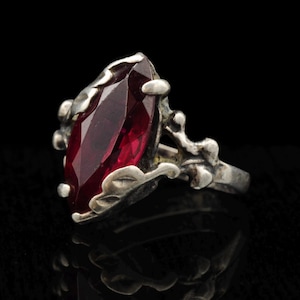 May include: A silver ring featuring a large, marquise-cut, deep red gemstone set within a textured, organic-looking metal band. The metalwork surrounding the stone has a leaf-like pattern with raised prongs that secure the faceted red center stone.