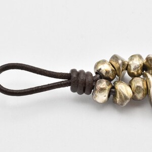 K&ouml;nnte beinhalten: Nahaufnahme eines Armbands mit einer Schlaufe aus dunkelbraunem Lederband und einer Reihe unregelm&auml;&szlig;ig geformter, goldfarbener Metallperlen. Die Perlen sind aneinandergereiht und bilden ein einzigartiges und strukturiertes Design. Das Armband befindet sich vor einem schlichten wei&szlig;en Hintergrund.