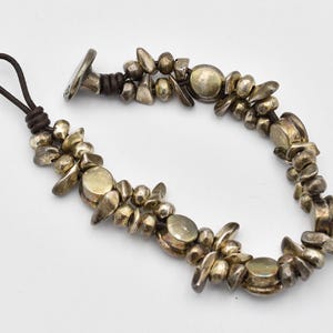 K&ouml;nnte beinhalten: Ein silberfarbenes Armband mit einer dunkelbraunen Lederschnur. Das Armband hat unregelm&auml;&szlig;ig geformte Metallperlen und kreisf&ouml;rmige Akzente. Der Verschluss ist ein silberfarbener Knebelverschluss. Das Armband liegt auf wei&szlig;em Grund.