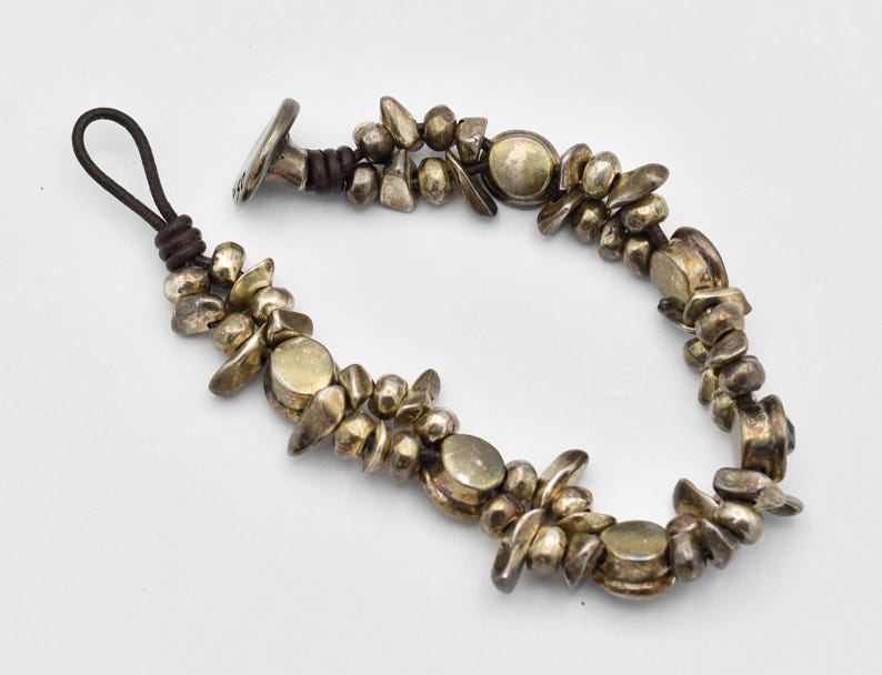 K&ouml;nnte beinhalten: Ein silberfarbenes Armband mit einer dunkelbraunen Lederschnur. Das Armband hat unregelm&auml;&szlig;ig geformte Metallperlen und kreisf&ouml;rmige Akzente. Der Verschluss ist ein silberfarbener Knebelverschluss. Das Armband liegt auf wei&szlig;em Grund.