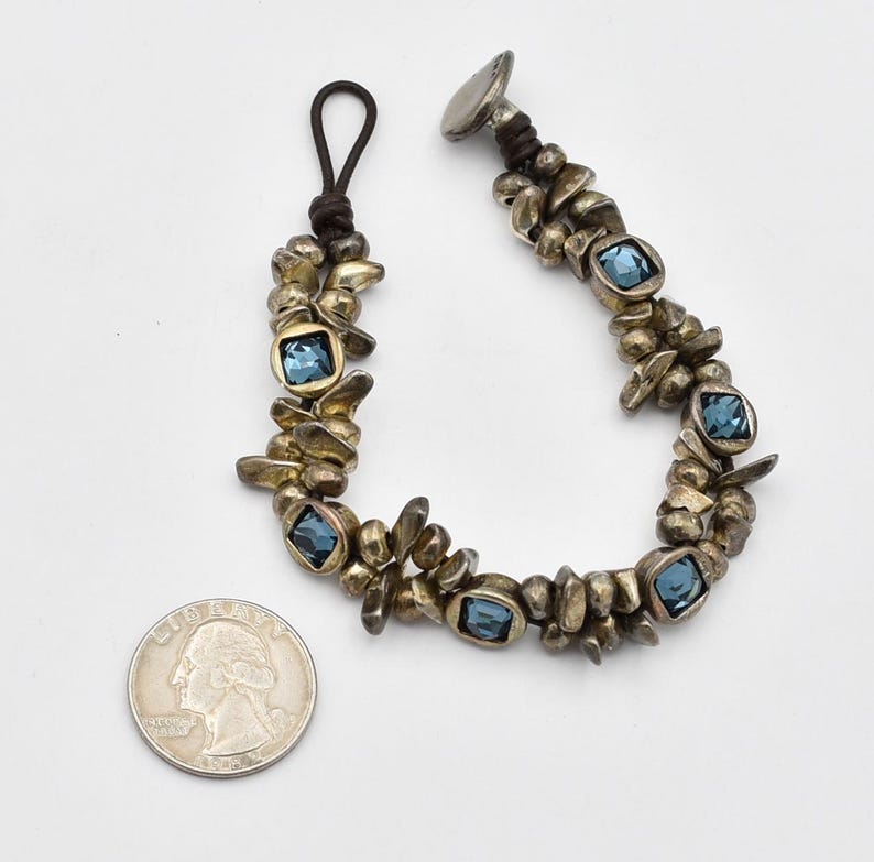 K&ouml;nnte beinhalten: Ein silberfarbenes Armband mit blauen quadratischen Edelsteinen. Das Armband hat ein einzigartiges Design mit unregelm&auml;&szlig;igen Metallformen und einem Lederkordelverschluss. Eine M&uuml;nze ist als Gr&ouml;&szlig;enreferenz enthalten.
