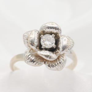 May include: A silver-toned ring featuring a sculpted flower design with textured petals. A single, clear, round-cut gemstone is prong-set in the center of the flower, serving as the focal point of the piece. The band is smooth and metallic.