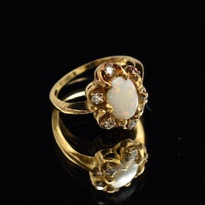 May include: A gold ring featuring a central oval-shaped opal gemstone with an iridescent play of color. The opal is surrounded by a scalloped gold setting accented with small, round, clear stones embedded within the metal frame of the ring.