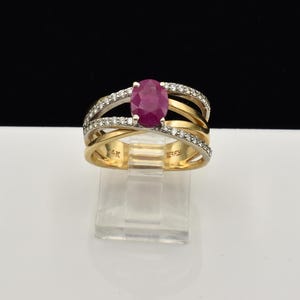 May include: A 14K gold ring featuring a central oval-cut deep pink ruby gemstone. The band is designed with overlapping, intertwined gold strands, with two of the strands accented by small, clear pavé-set diamonds. The interior is stamped 14K.