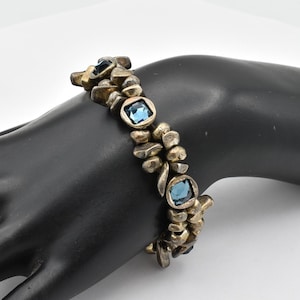 K&ouml;nnte beinhalten: Ein Vintage-Armband mit einzigartigem Design. Das Armband verf&uuml;gt &uuml;ber unregelm&auml;&szlig;ig geformte, goldfarbene Metallglieder und quadratische, hellblaue Edelsteine. Die Edelsteine sind in einem quadratischen Rahmen gefasst. Das Armband wird auf einer schwarzen Oberfl&auml;che pr&auml;sentiert.