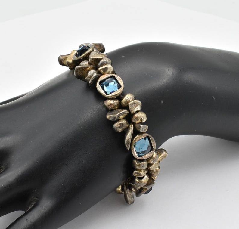 K&ouml;nnte beinhalten: Ein Vintage-Armband mit einzigartigem Design. Das Armband verf&uuml;gt &uuml;ber unregelm&auml;&szlig;ig geformte, goldfarbene Metallglieder und quadratische, hellblaue Edelsteine. Die Edelsteine sind in einem quadratischen Rahmen gefasst. Das Armband wird auf einer schwarzen Oberfl&auml;che pr&auml;sentiert.