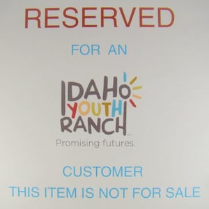 May include: A white sign with the word "RESERVED" in red at the top. Below, the text "FOR AN" is in blue. The Idaho Youth Ranch logo is in the center, and the text "CUSTOMER" and "THIS ITEM IS NOT FOR SALE" are at the bottom.