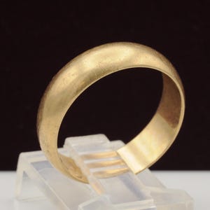 May include: A solid gold band ring with a smooth, rounded exterior surface and a polished finish. The ring features a simple, continuous circular design with visible fine surface scratches consistent with wear, displayed on a clear stand.