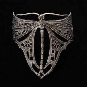 Puede incluir: Un brazalete de metal en tono plateado con un diseño detallado de libélula. Las alas están elaboradas con un intrincado patrón de red calado que se extiende por la banda, mientras que el cuerpo de la libélula forma el elemento central vertical.