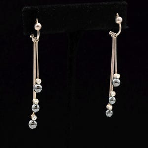May include: A pair of silver-toned drop earrings featuring two thin vertical bars suspended from a ball-post backing. Each earring is accented with three small, dark gray spherical beads and three clear, round faceted stones arranged in an alternating pattern.
