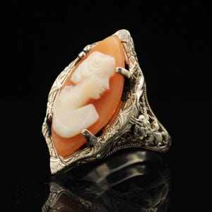 May include: A marquise-shaped ring featuring a carved cameo of a profile portrait in cream-colored material set against an orange background. The setting is made of silver-toned metal with intricate floral engravings and filigree detailing.