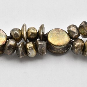 K&ouml;nnte beinhalten: Nahaufnahme einer Halskette mit unregelm&auml;&szlig;ig geformten, silberfarbenen Perlen. Die Perlen sind auf einer dunklen Schnur aufgereiht, wobei einige flacher und andere runder erscheinen. Die Halskette hat eine Vintage- oder handwerkliche &Auml;sthetik.