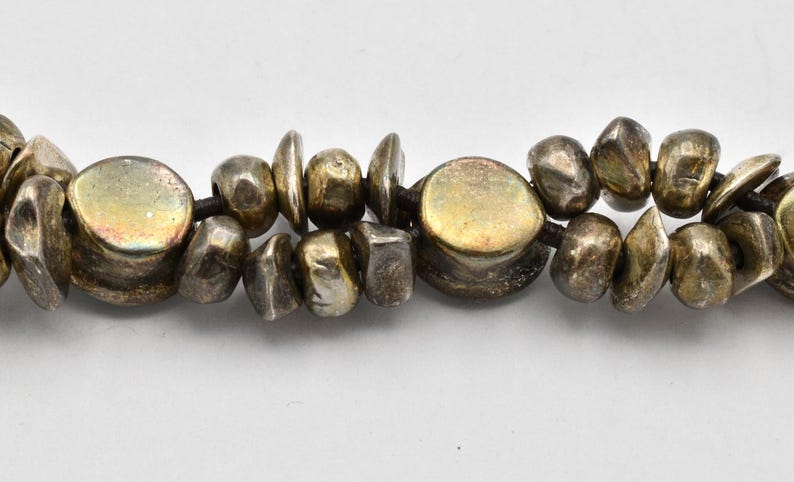 K&ouml;nnte beinhalten: Nahaufnahme einer Halskette mit unregelm&auml;&szlig;ig geformten, silberfarbenen Perlen. Die Perlen sind auf einer dunklen Schnur aufgereiht, wobei einige flacher und andere runder erscheinen. Die Halskette hat eine Vintage- oder handwerkliche &Auml;sthetik.