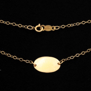 May include: A gold chain necklace featuring a smooth, polished oval-shaped pendant centered on the chain. The necklace includes a circular spring ring clasp and a small metal tag near the closure with visible engraved text.