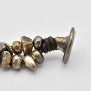 K&ouml;nnte beinhalten: Nahaufnahme eines Armbands mit unregelm&auml;&szlig;ig geformten, silberfarbenen Perlen und einer dunkelbraunen Lederschnur. Das Armband hat einen silberfarbenen Verschluss. Die Perlen haben ein abgenutztes, antikes Aussehen.