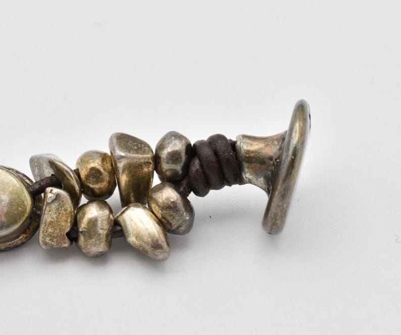 K&ouml;nnte beinhalten: Nahaufnahme eines Armbands mit unregelm&auml;&szlig;ig geformten, silberfarbenen Perlen und einer dunkelbraunen Lederschnur. Das Armband hat einen silberfarbenen Verschluss. Die Perlen haben ein abgenutztes, antikes Aussehen.