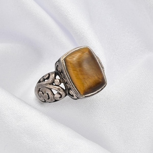 May include: A silver ring with a rectangular tiger's eye stone. The stone is a warm brown color with a golden sheen. The ring has an ornate, openwork design on the band.