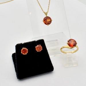 May include: A jewelry set consisting of a gold necklace with a round orange gemstone pendant, a gold ring featuring a matching round orange gemstone, and a pair of gold stud earrings with round orange gemstones set on a black velvet display.