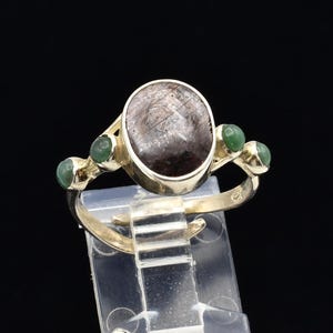 May include: A silver ring featuring a large, oval, dark brown gemstone with a web-like pattern. The ring is accented with three small, round, green gemstones on each side of the main stone. The ring is displayed on a clear stand against a black background.
