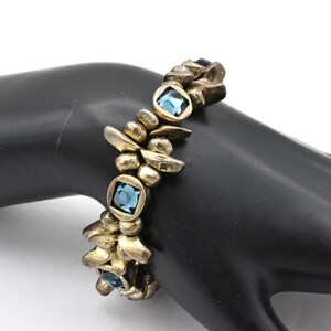 K&ouml;nnte beinhalten: Ein goldfarbenes Armband mit quadratischen, hellblauen Edelsteinen, die in einem strukturierten, organischen Design gefasst sind. Das Armband besteht aus miteinander verbundenen, unregelm&auml;&szlig;igen Formen und ist ein einzigartiges Accessoire. Das Armband wird auf einer schwarzen Oberfl&auml;che pr&auml;sentiert.