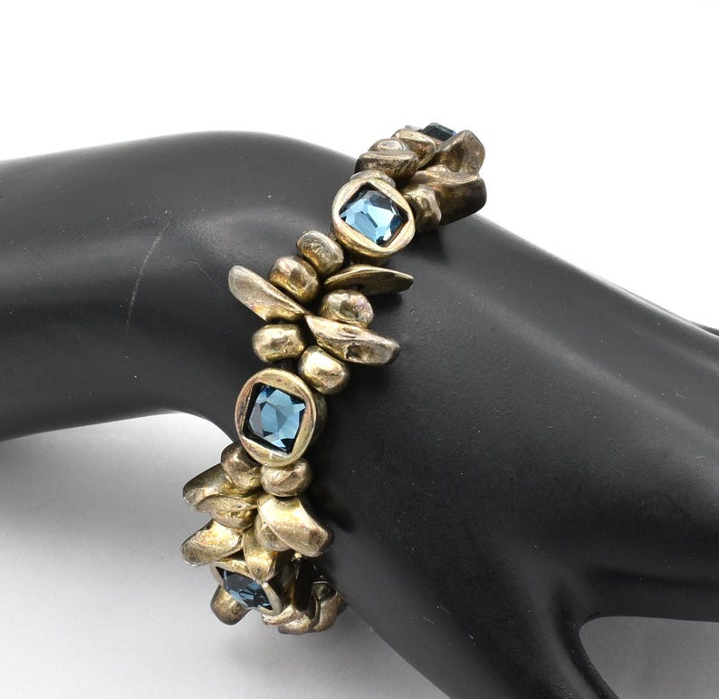 K&ouml;nnte beinhalten: Ein goldfarbenes Armband mit quadratischen, hellblauen Edelsteinen, die in einem strukturierten, organischen Design gefasst sind. Das Armband besteht aus miteinander verbundenen, unregelm&auml;&szlig;igen Formen und ist ein einzigartiges Accessoire. Das Armband wird auf einer schwarzen Oberfl&auml;che pr&auml;sentiert.