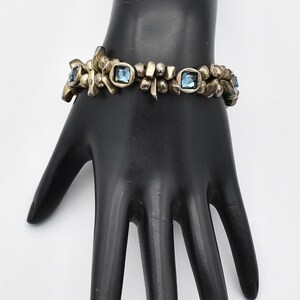 K&ouml;nnte beinhalten: Vintage-Armband mit floralem Design und quadratischen blauen Edelsteinen. Das Armband besteht aus goldfarbenem Metall und wird auf einer schwarzen Handmannequin pr&auml;sentiert. Ein einzigartiges Schmuckst&uuml;ck.