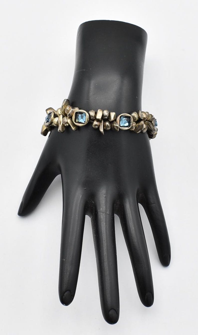 K&ouml;nnte beinhalten: Vintage-Armband mit floralem Design und quadratischen blauen Edelsteinen. Das Armband besteht aus goldfarbenem Metall und wird auf einer schwarzen Handmannequin pr&auml;sentiert. Ein einzigartiges Schmuckst&uuml;ck.