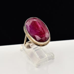 May include: A ring featuring a large, oval-shaped, faceted deep red gemstone set in a polished silver-toned metal band. The stone displays a rich, saturated crimson hue with visible internal facets that catch the light, mounted in a simple bezel setting.