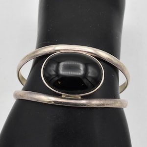 May include: A silver cuff bracelet with a large, oval black stone at the center. The bracelet has a simple, elegant design with a polished finish. The black stone is smooth and reflective, set between the curved silver bands.