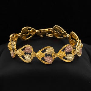 Op de afbeelding: Een driekleurige gouden armband met een herhalend patroon van hartvormige schakels, elk voorzien van gestructureerde bladmotieven in geel-, rosé- en witgoud. De armband is voorzien van een metalen sluiting en heeft een gepolijste afwerking.