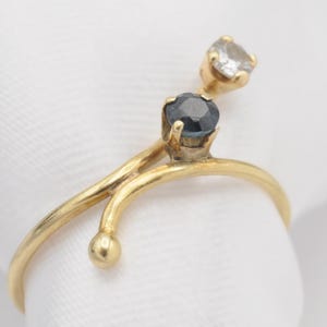 May include: A gold-toned ring with a unique design. The ring features a dark blue gemstone and a clear gemstone, both set on a curved band. The ring's design includes a small gold ball. The ring is displayed on a white surface.