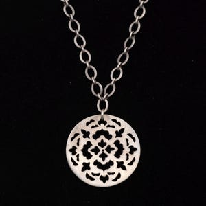 May include: Silver necklace with a circular pendant. The pendant has a detailed, openwork floral design. The necklace hangs from a linked chain against a black background. This is a piece of jewellery.