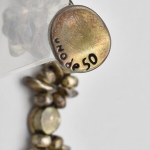 K&ouml;nnte beinhalten: Nahaufnahme eines goldfarbenen Anh&auml;ngers mit der Inschrift "UNO de 50" in Schwarz. Der Anh&auml;nger ist an einer Perlenkette befestigt. Die Kette und der Anh&auml;nger haben ein Vintage-Aussehen.