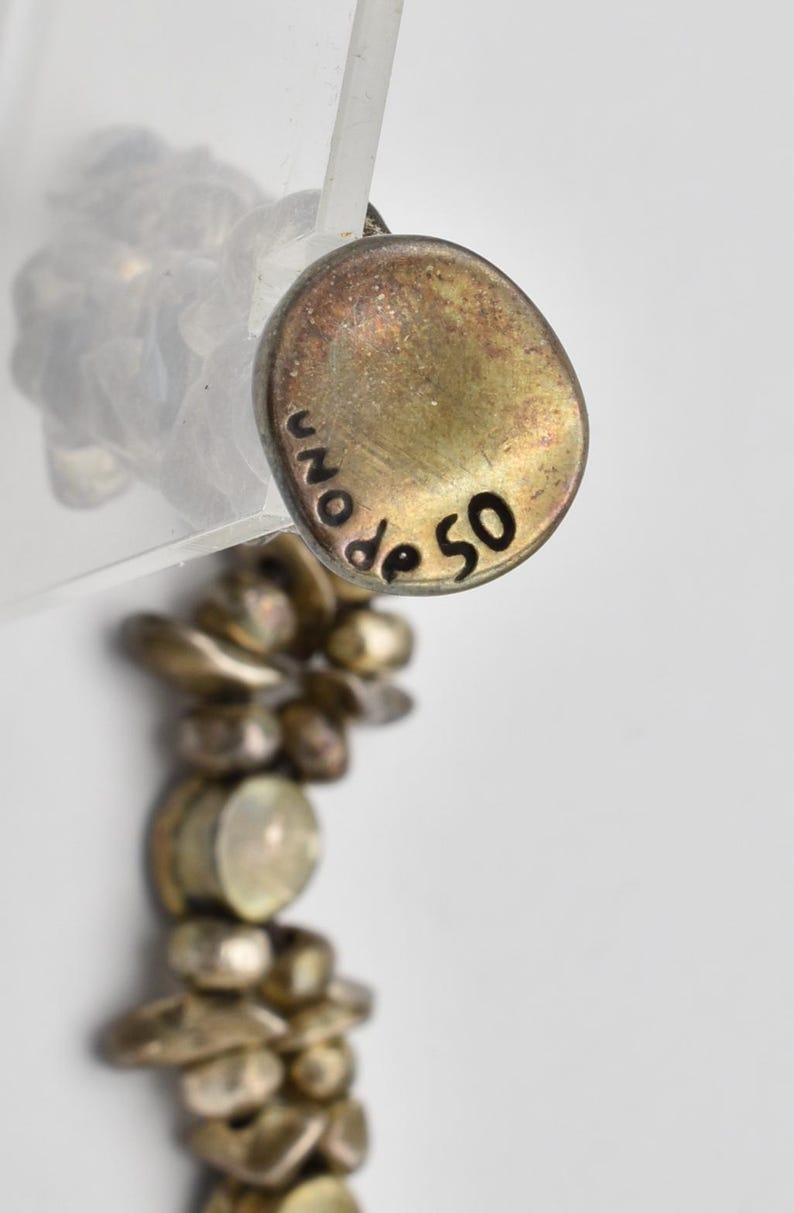 K&ouml;nnte beinhalten: Nahaufnahme eines goldfarbenen Anh&auml;ngers mit der Inschrift "UNO de 50" in Schwarz. Der Anh&auml;nger ist an einer Perlenkette befestigt. Die Kette und der Anh&auml;nger haben ein Vintage-Aussehen.