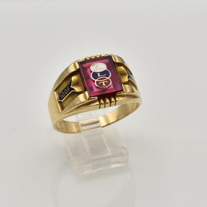 May include: A gold ring featuring a rectangular red stone set in the centre. The stone displays three interlocking circles with the letters F, L, and T. The side of the ring band is engraved with the black text I.O.O.F. on a dark triangular background.