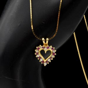 May include: A gold necklace featuring a heart-shaped pendant. The pendant has a central black heart cutout surrounded by a border of alternating small round red rubies and clear white diamonds set in a gold frame on a thin gold chain.