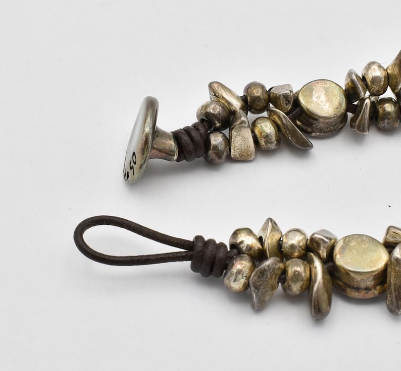 K&ouml;nnte beinhalten: Nahaufnahme eines silberfarbenen Armbands mit Lederband. Das Armband hat verschiedene Formen von Silberperlen und einen Verschluss mit der eingravierten Zahl "50". Das Lederband ist dunkelbraun.