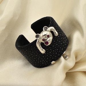 May include: A black cuff bracelet with a textured surface and a silver bear design. The bear has red gemstone eyes and is attached to the bracelet. The bracelet is a unique piece of jewelry.