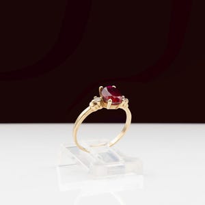 May include: A gold ring with a large, oval-cut ruby as the central stone. The ring is flanked by smaller, clear gemstones. The ring is presented on a clear plastic stand, set against a dark backdrop. The ring's design is classic and elegant.
