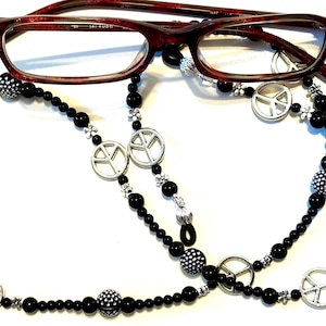 May include: Black beaded eyeglass chain with silver peace sign charms. The chain is worn with red-framed glasses.