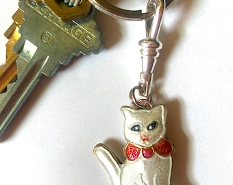 May include: A white enamel cat charm with red and gold accents on a silver key chain. The cat is wearing a red and gold collar.