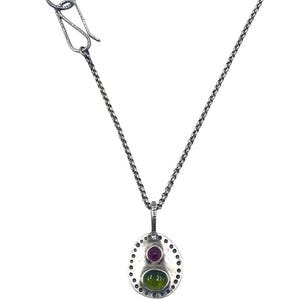 May include: A silver-toned necklace with a pendant featuring a red and green gemstone. The pendant is oval-shaped with a dotted border. The necklace chain has a textured design and a hook clasp.