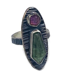 May include: A silver ring with an oval face featuring a small, pink, hexagonal gemstone and a larger, green, geometric gemstone. The ring has a textured, etched design on the face and band.